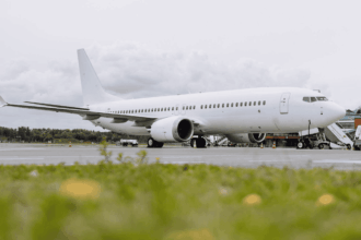 Side view of a white, unbranded narrow‑body passenger aircraft parked on an airport apron, photographed from a low angle across a strip of green grass in the foreground. The aircraft’s fuselage, wings and tail are clean and undecorated, with engines mounted beneath the wings and the landing gear resting on the concrete surface. Ground service equipment and mobile stairs are positioned near the forward section, while low airport buildings and trees sit in the background. The scene is lit by soft daylight under a grey, cloud‑filled sky, giving the image a muted, industrial atmosphere.