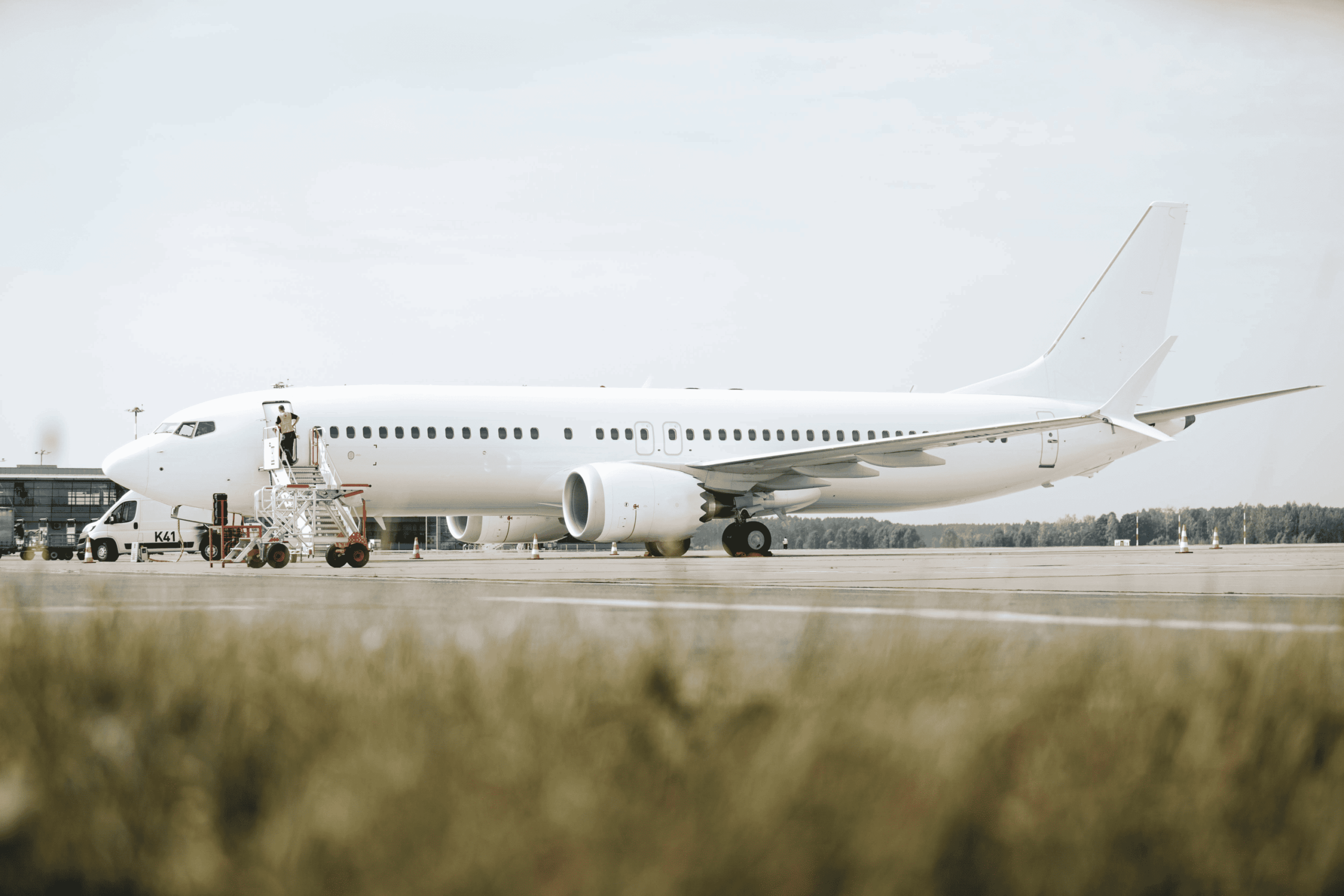 Side view of an all‑white narrow‑body passenger jet parked on an airport apron, with no visible airline branding on the fuselage or tail. A mobile stairway is positioned at the forward left door, where a person stands at the top of the steps, while a small service vehicle is parked alongside. The aircraft’s engines hang beneath the wings, and the landing gear rests on the concrete surface marked with faint lines and cones. In the foreground, out‑of‑focus grass frames the lower edge of the image, while low airport buildings and a tree line sit in the distance under a pale, lightly overcast sky.