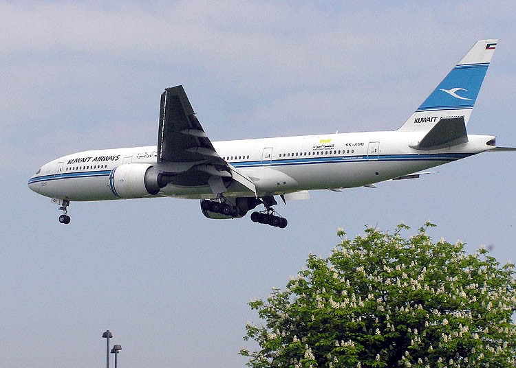 A Kuwait Airways Boeing 777-200 (registration 9K-AOB) the aircraft is shown with landing gear deployed, flying low over greenery against an overcast sky.