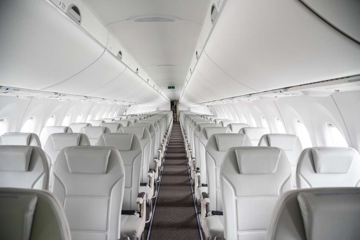 Interior of Airbus A220-300 airBaltic cabin taken from the front end of the aisle. The seats are white placed in a 3-2 configurationall the way along, with overhead storage visible above each row. Windows can be seen adorning either side.