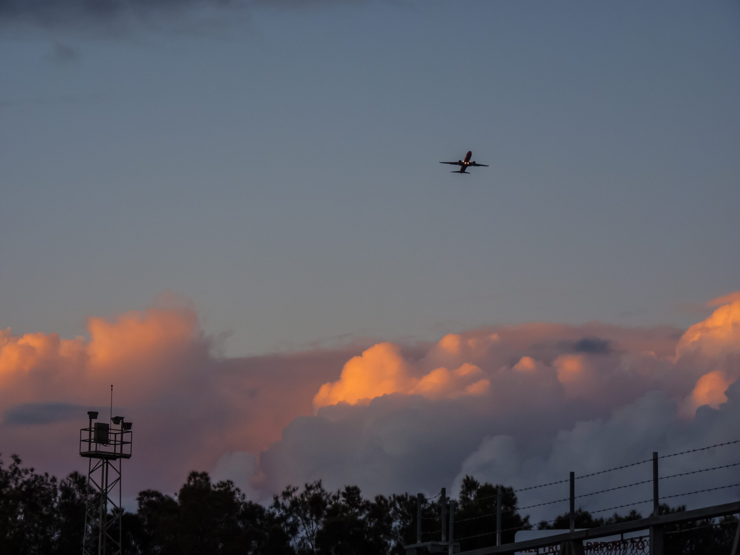 Distant view of a small passenger aircraft flying across a wide evening sky, silhouetted against soft blue and grey tones. Large, billowing clouds glow with warm pink and orange light from the setting sun near the horizon. Along the bottom edge of the image, dark silhouettes of trees, fencing and a metal tower with mounted equipment frame the scene, emphasising the aircraft’s height and the expansive sky above.