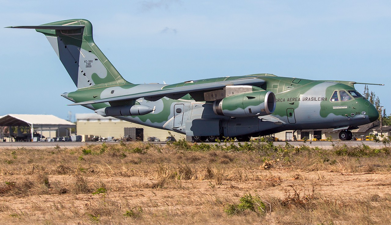 The image shows a KC-390 Millennium aircraft out of flight. The aircraft has landed atop yellowing dessert grass. The sky is blue in the background.