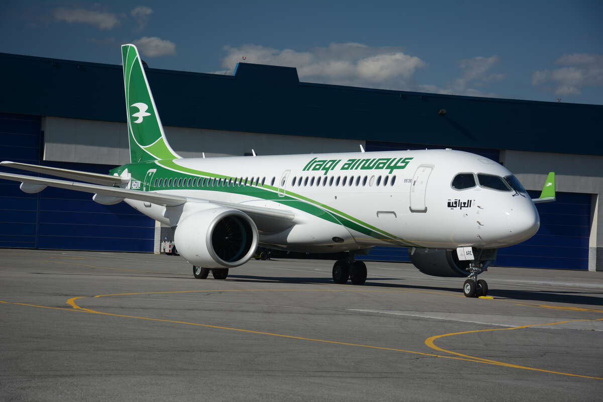 An Airbus A220 aircraft with the green Iraqi Airways logo is in a taxiway, positioned in the centre of the image. It is facing towards the camera, with the nose of the aircraft centre right of the image. The sky is blue in the background, and the ground is tarmac in the foreground.