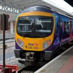 The image depicts thefront of a TransPennine class 185 train as it prepares to leave Manchester Airport Station. The front is yellow, and the body of the train is dark blue, light blue, and purple. The Front of the train is the main focus of the image, and is central.