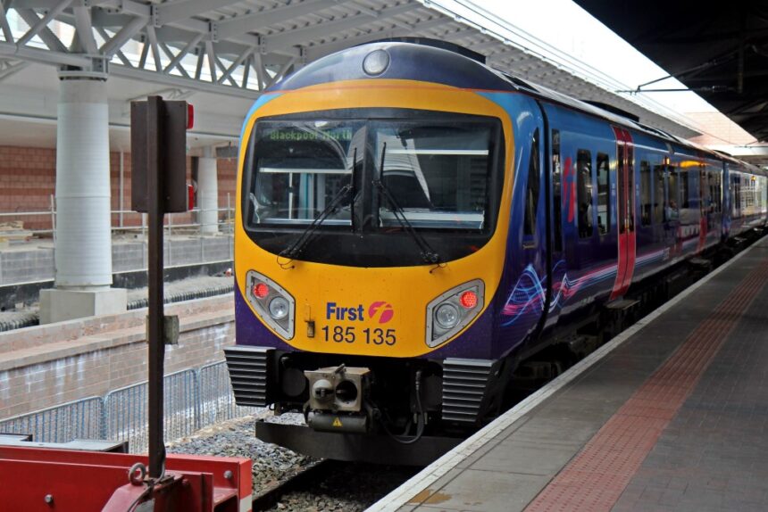 The image depicts thefront of a TransPennine class 185 train as it prepares to leave Manchester Airport Station. The front is yellow, and the body of the train is dark blue, light blue, and purple. The Front of the train is the main focus of the image, and is central.