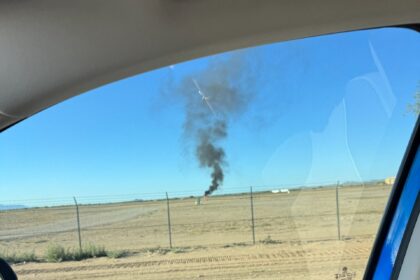 Shot from inside car, looking out of the window to see a field with an aircraft on fire, black smoke in the air.