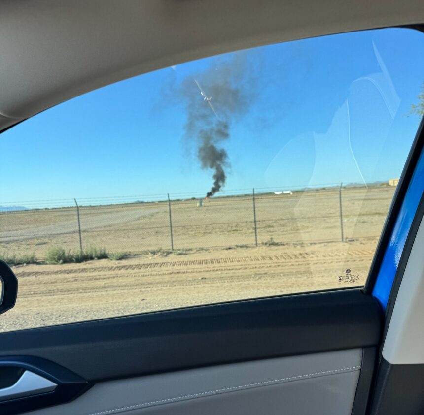 Shot from inside car, looking out of the window to see a field with an aircraft on fire, black smoke in the air.