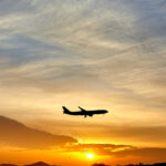 Shadowed plane in the centre flying through a pale blue, orange and yellow sunset in the distance. Plane is facing to the right.