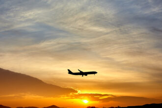 Shadowed plane in the centre flying through a pale blue, orange and yellow sunset in the distance. Plane is facing to the right.