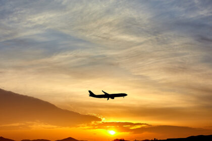 Shadowed plane in the centre flying through a pale blue, orange and yellow sunset in the distance. Plane is facing to the right.