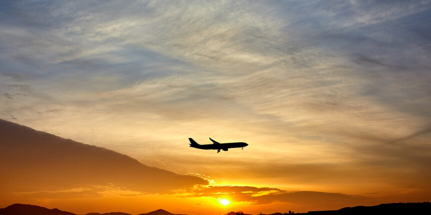 Shadowed plane in the centre flying through a pale blue, orange and yellow sunset in the distance. Plane is facing to the right.