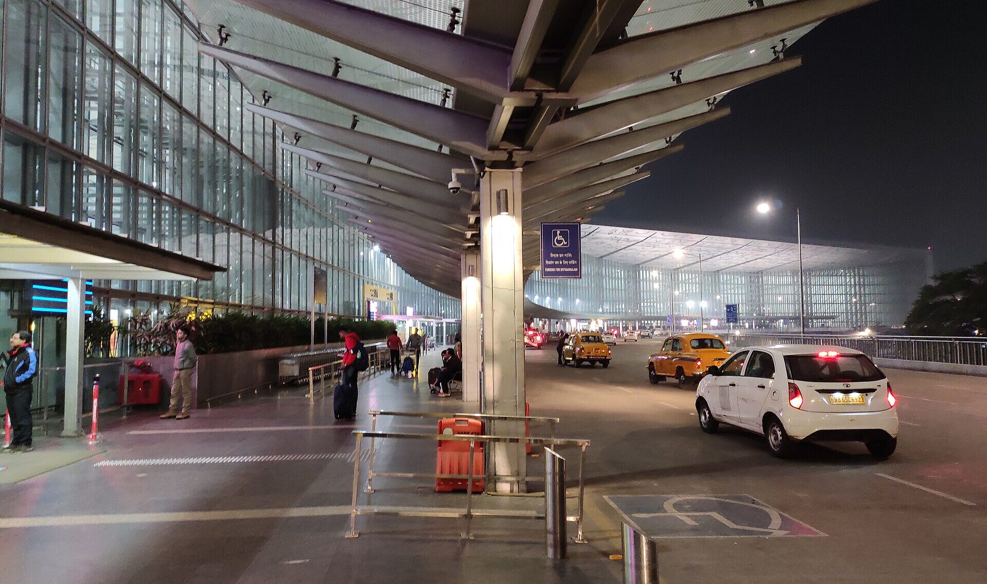 Exterior of Netaji Subhash Chandra Bose International Airport at night, taxis waiting outside, people with their luggage.