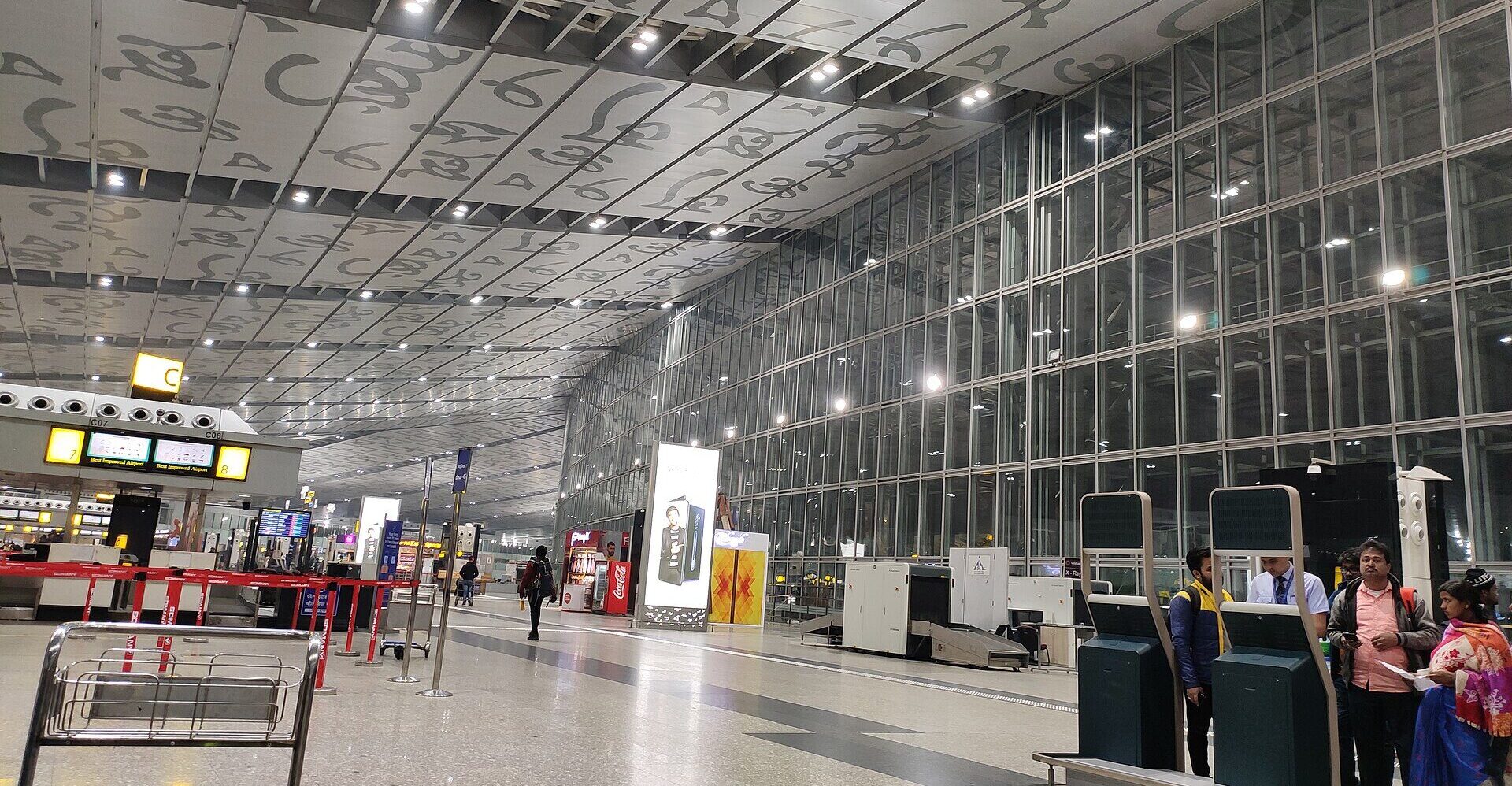 Interior of Netaji Subhas Chandra Bose International Airport, symbols written on the ceiling, glass panels on the sides, people waiting with luggage.