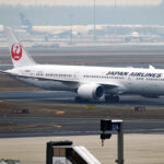 A Japan Airlines plane on an airport runway. The plane is white with 'Japan Airlines' in black lettering across the front end of it, and has the red Japan Airlines logo on the tail. The plane is positioned to the centre right of the image, and the remaining background is desert-looking.