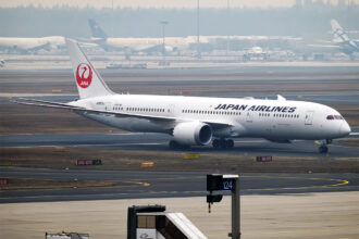 A Japan Airlines plane on an airport runway. The plane is white with 'Japan Airlines' in black lettering across the front end of it, and has the red Japan Airlines logo on the tail. The plane is positioned to the centre right of the image, and the remaining background is desert-looking.