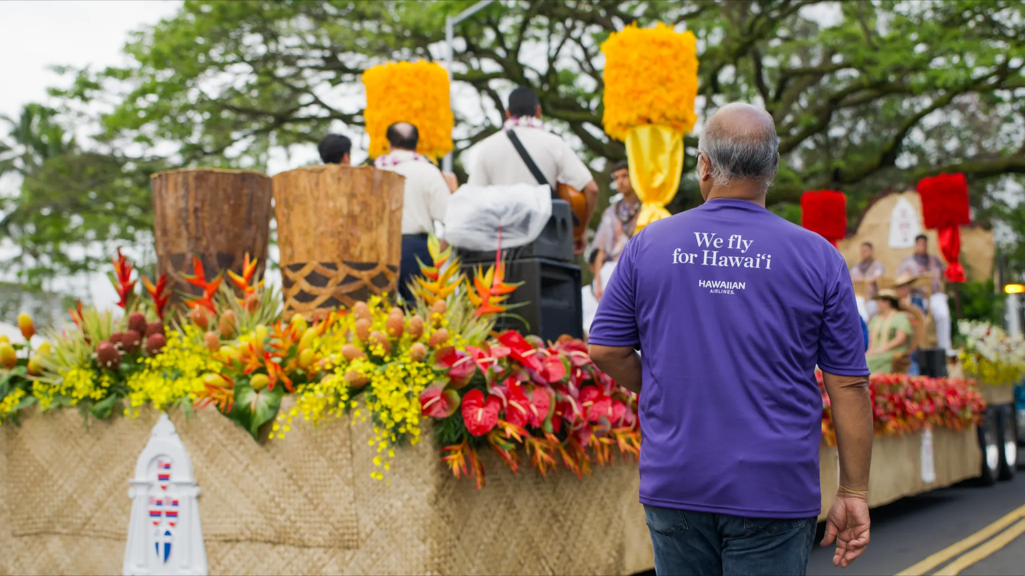 Man in a purple shirt that says "we fly for Hawai'i". Stage in the background, with people, bright flowers and trees.