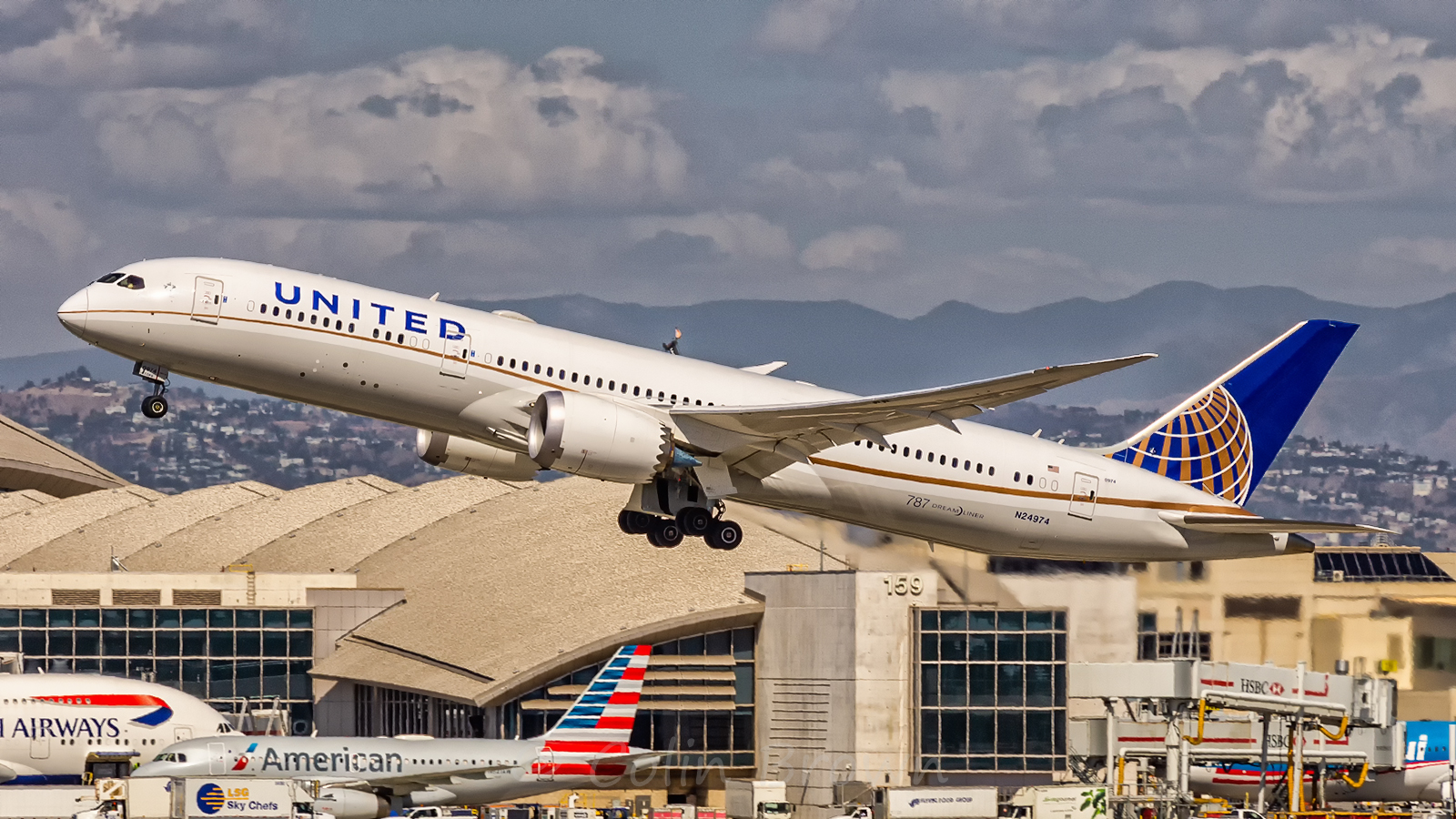 A Boeing 787-9 Dreamliner, United Airlines, taking off, with buildings in the background. Other aircraft on the apron, grey sky in the background.