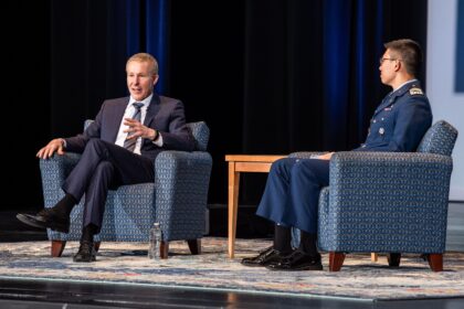 Two men seated on separate blue upholstered armchairs on a stage, facing slightly towards each other. One person wears a dark business suit, crosses their legs, and gestures with one hand, while the other wears a dark formal uniform with insignia and sits upright. A small wooden side table stands between the chairs, with a water bottle on the floor nearby. The stage is lit from above, with a patterned rug underfoot and a dark backdrop with vertical blue lighting panels behind the men.