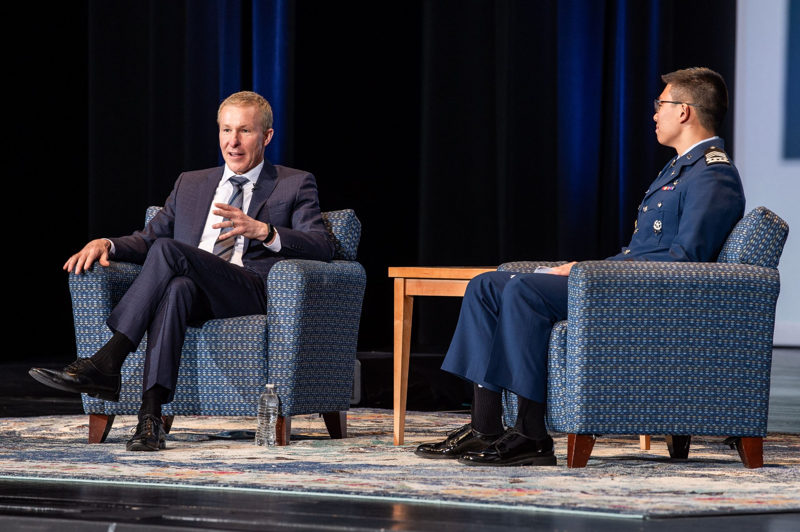 Two men seated on separate blue upholstered armchairs on a stage, facing slightly towards each other. One person wears a dark business suit, crosses their legs, and gestures with one hand, while the other wears a dark formal uniform with insignia and sits upright. A small wooden side table stands between the chairs, with a water bottle on the floor nearby. The stage is lit from above, with a patterned rug underfoot and a dark backdrop with vertical blue lighting panels behind the men.