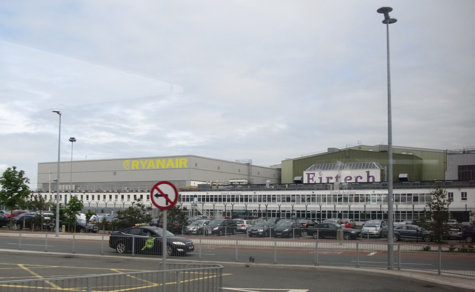 Wide view of aircraft maintenance hangars at Dublin Airport, with large industrial buildings in the background displaying the yellow Ryanair logo and the Eirtech sign. A long, low office block and a busy car park filled with parked vehicles sit in front of the hangars. In the foreground, a road runs past the airport perimeter fencing, with a “no right turn” traffic sign, streetlights and a black car passing by under a pale, overcast sky.