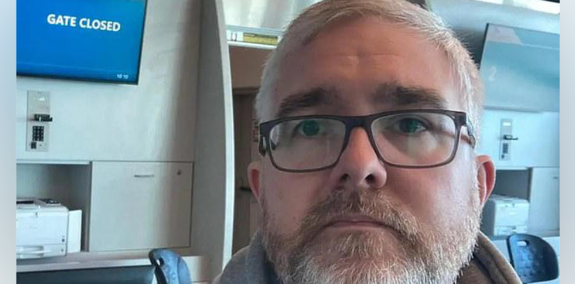 A man with glasses and grey hair sitting in front of a sign reading 'Gate closed'