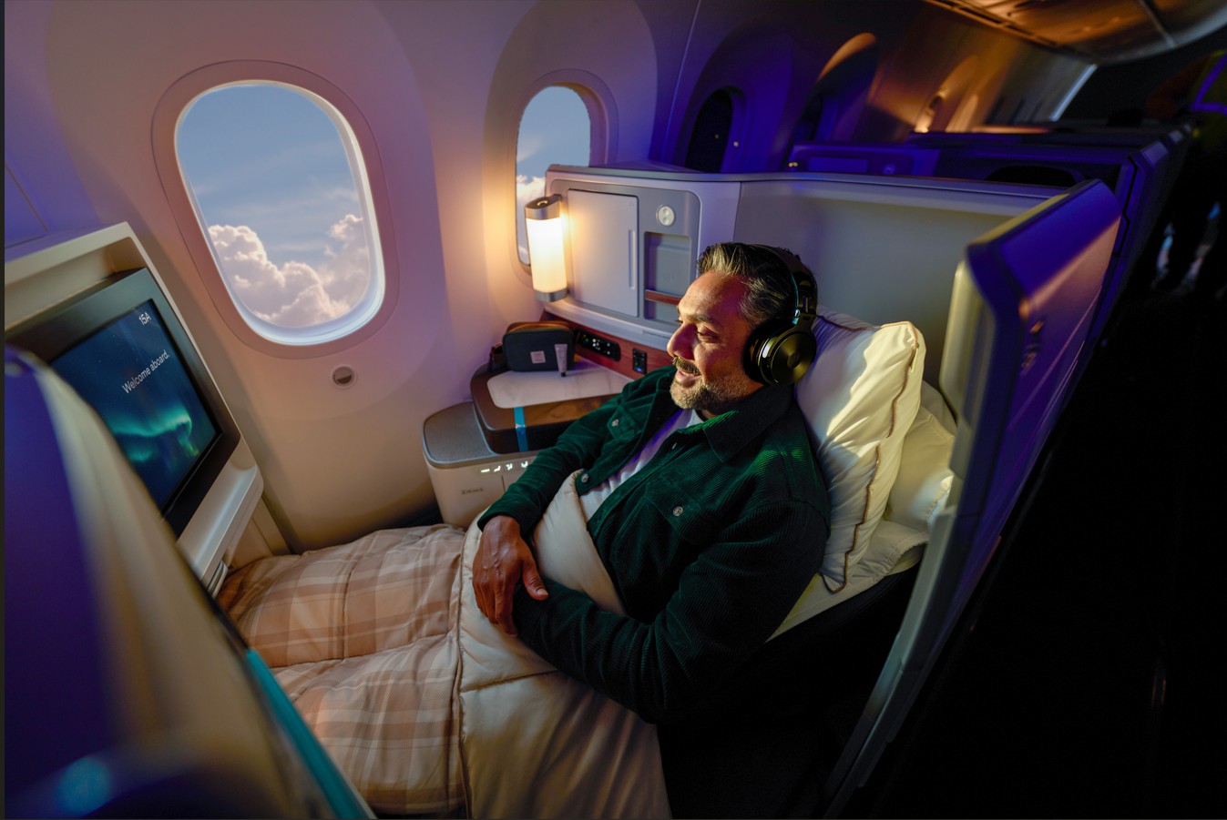 A passenger relaxing in a business-class airplane seat that has been converted into a fully flat bed, lying on his back with a blanket pulled up to his waist.