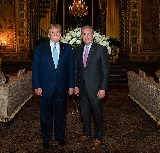 Two men wearing suits standing inside a mansion with a chandelier.
