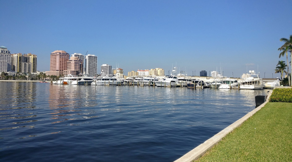A waterfront with white boats and tall buildings behind it under a blue sky.