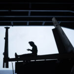 Silhouetted view inside an aircraft hangar, showing a person seated on the horizontal tail section of an aircraft, high above the ground. Large vertical tail fins frame the scene, with a ladder leaning up from the left. The open hangar door reveals a pale sky and a faint horizon line over the sea, contrasting with the dark interior structure and overhead beams.