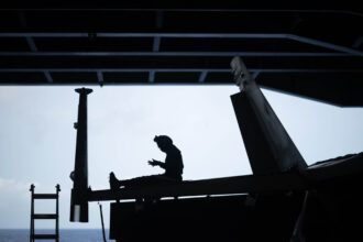 Silhouetted view inside an aircraft hangar, showing a person seated on the horizontal tail section of an aircraft, high above the ground. Large vertical tail fins frame the scene, with a ladder leaning up from the left. The open hangar door reveals a pale sky and a faint horizon line over the sea, contrasting with the dark interior structure and overhead beams.