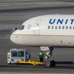 Close-up view of the front section of a United Airlines Boeing 757 on the airport apron, showing the white nose and cockpit windows with the blue “UNITED” lettering along the fuselage. The aircraft’s nose landing gear is connected to a yellow tow bar attached to a small white pushback tug, with a high‑visibility‑clad ground crew member standing nearby. Grey concrete taxiways with painted markings stretch across the background under bright daylight.