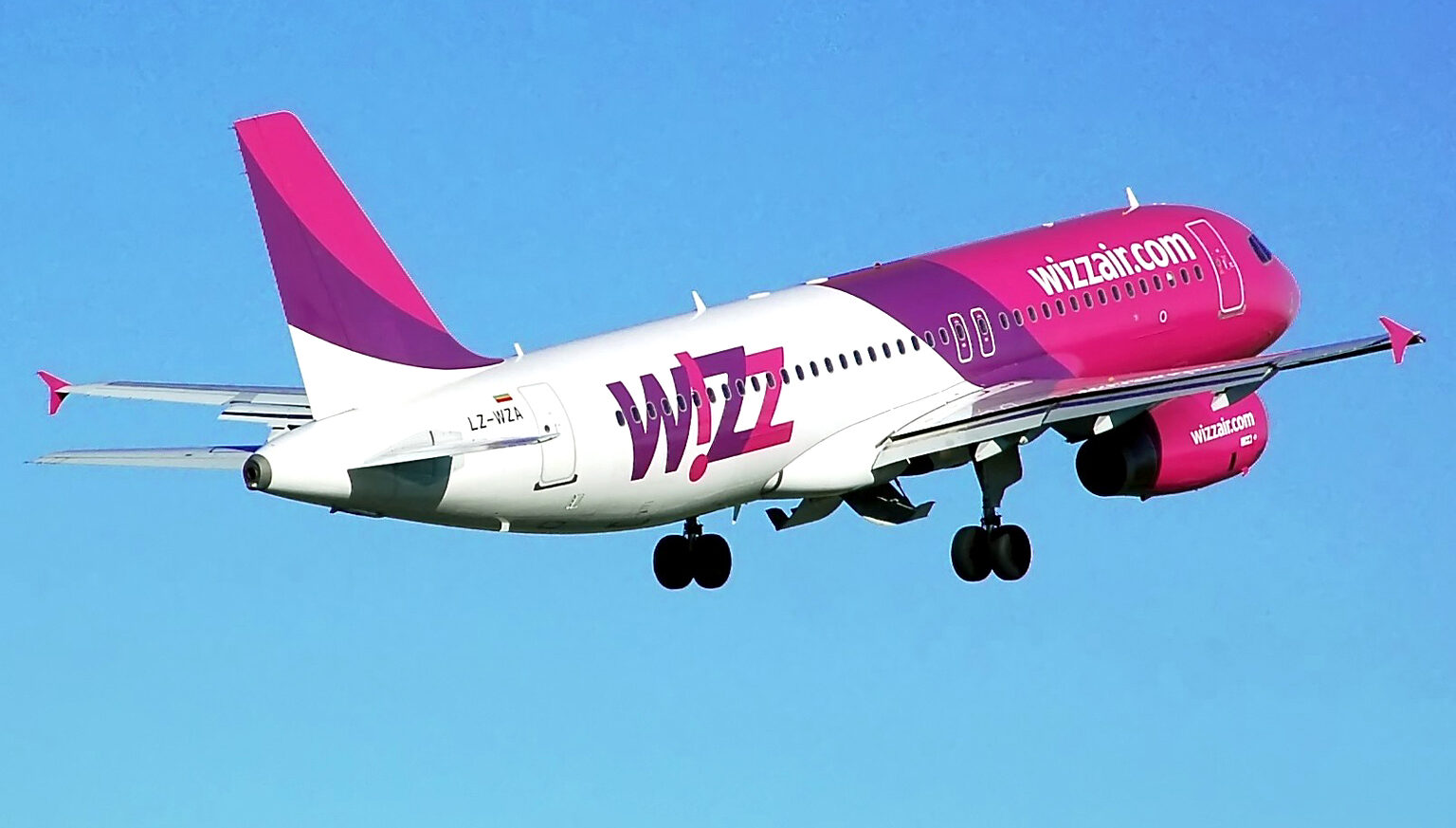 Wizz Air aircraft flying upwards into right top corner, through blue sky. Pink, purple and white livery.