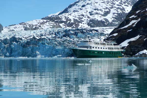 Two people in a red kayak paddling near a cruise ship in cold Alaskan waters with snowy mountains behind.
