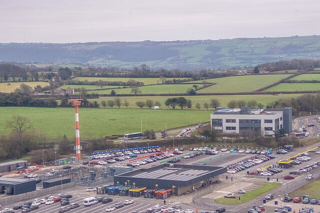 View from above of a corner of Bristol Airport car park, with two rectangular buildings in the low centre and mid right of the image. There are quite a few cars dotted around in parking spaces. There are green fields and trees in the background 