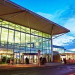 The front entrance to Bristol airport at dawn or dusk. The airport has long floor to ceiling windows across the front, a revolving style door extended out, and a flat white roof. People walk towards it.
