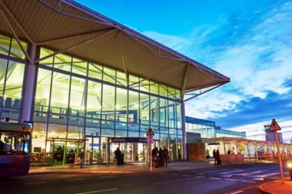 The front entrance to Bristol airport at dawn or dusk. The airport has long floor to ceiling windows across the front, a revolving style door extended out, and a flat white roof. People walk towards it.