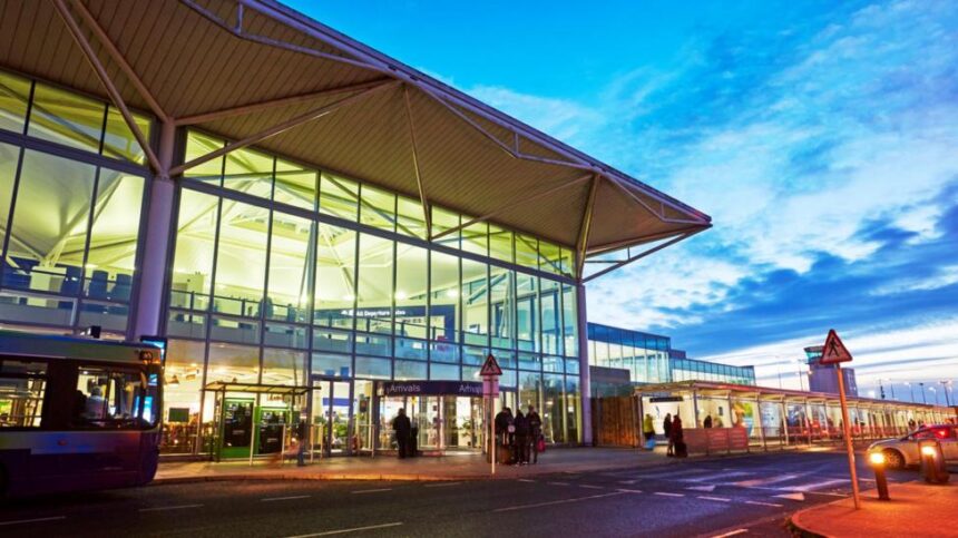 The front entrance to Bristol airport at dawn or dusk. The airport has long floor to ceiling windows across the front, a revolving style door extended out, and a flat white roof. People walk towards it.
