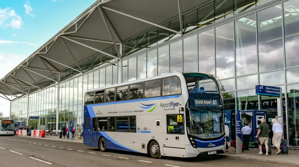 A blue and white double decker A1 bus is parked outside the front of Bristol Airport, which has a white roof and floor to ceiling windows. 