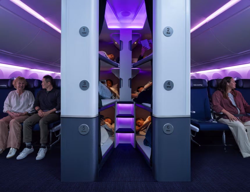 Image of the interior of the new Air New Zealand's Dreamliner, with purple lighting, people sat in seat and the Skynest area in the aisle between.