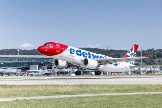 A Edelweiss Airbus A320 taking off; airport terminal in the background.
