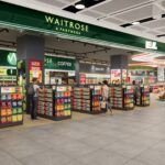 The image shows a dual branded Waitrose-Relay shop inside London Heathrow Airport Terminal 2. Along the top of the image is the dual branded logo in forest green and white. A range of colourful displays can be seen on stalls and shelves in the shop.