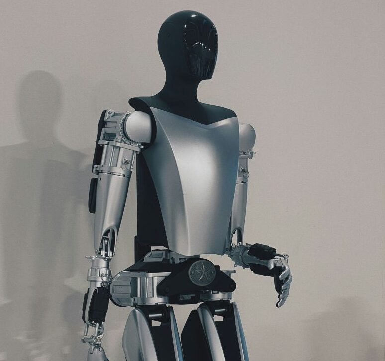 TA silver and black humanoid robot stands in the centre of the image, in front of a bare wall. The picture cuts off just below the robot's groin area. 
