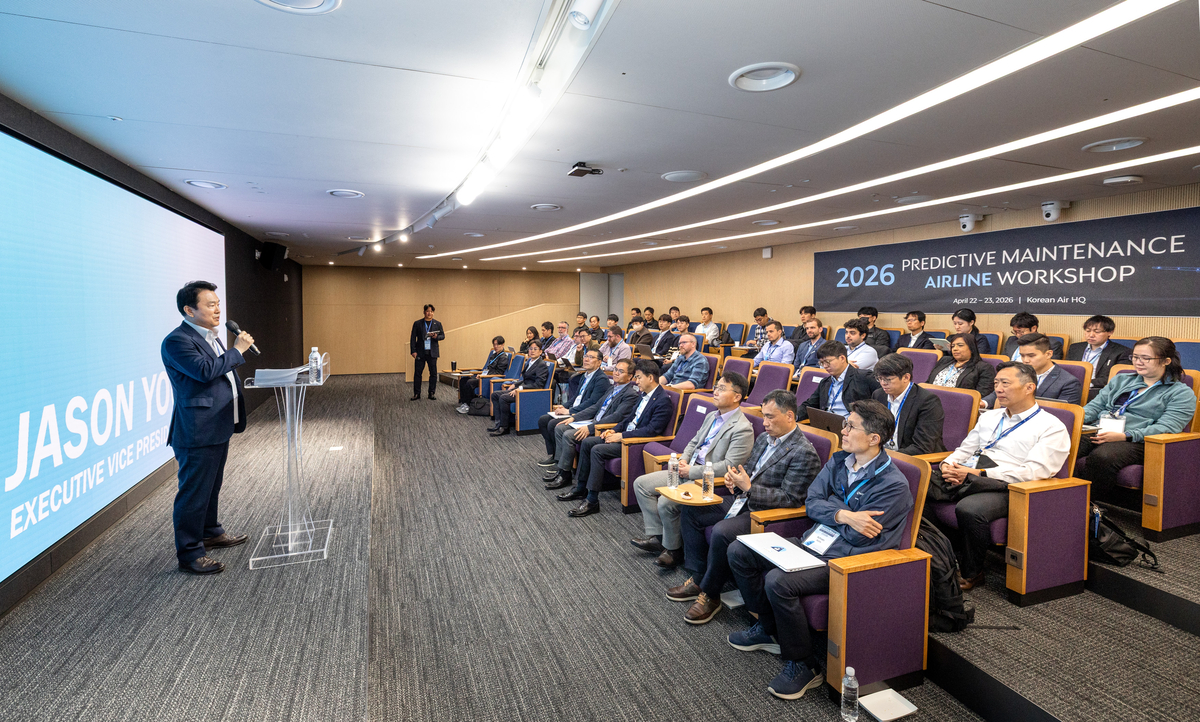 Image of a large meeting room with a person standing at the front giving a speech and many in seats watching.