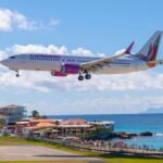 A Caribbean Airlines flight landing amid blue skies with a few clouds, some buildings surrounding, and clear blue sea water.