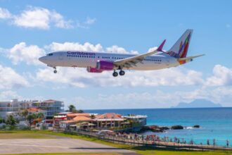 A Caribbean Airlines flight landing amid blue skies with a few clouds, some buildings surrounding, and clear blue sea water.