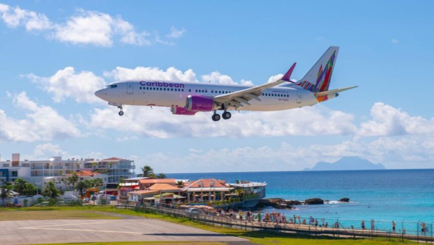 A Caribbean Airlines flight landing amid blue skies with a few clouds, some buildings surrounding, and clear blue sea water.