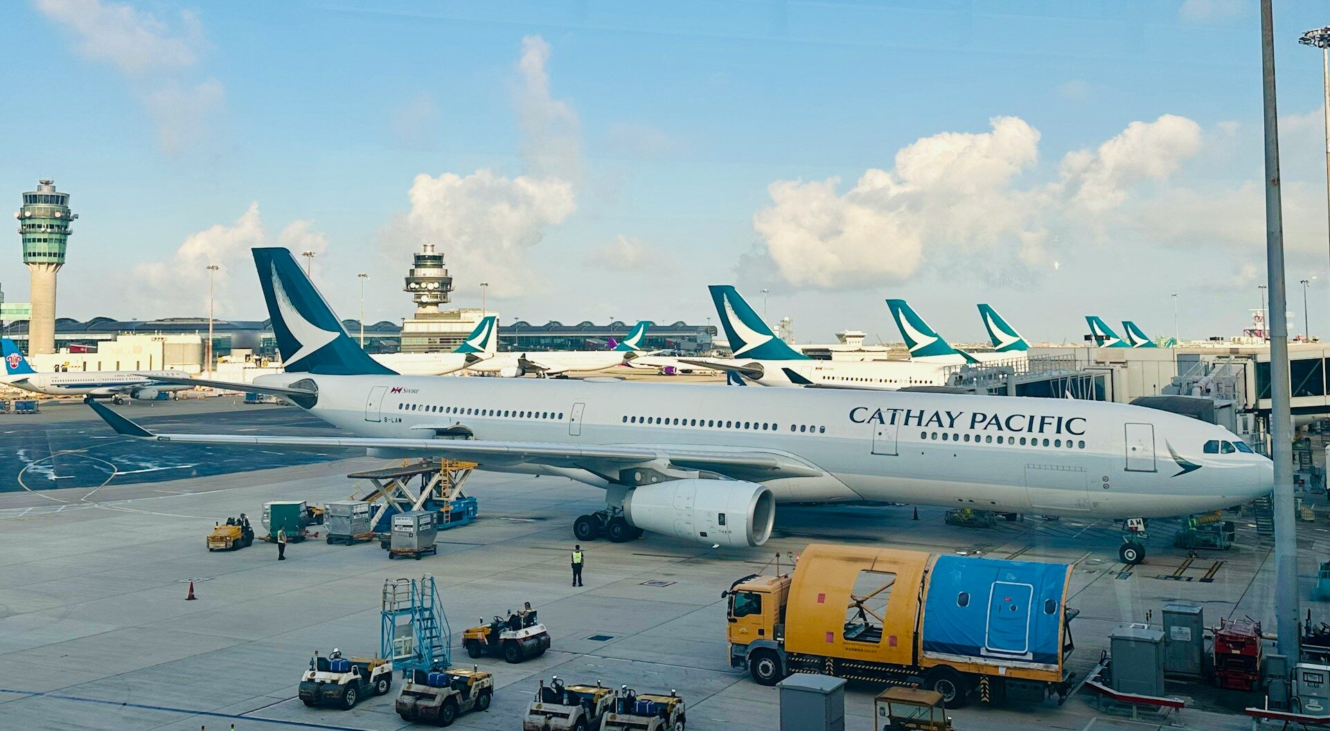 Cathay Pacific aircraft at Hong Kong International Airport, parked in apron. Ground vehicles around the aircraft, blue sky in background.