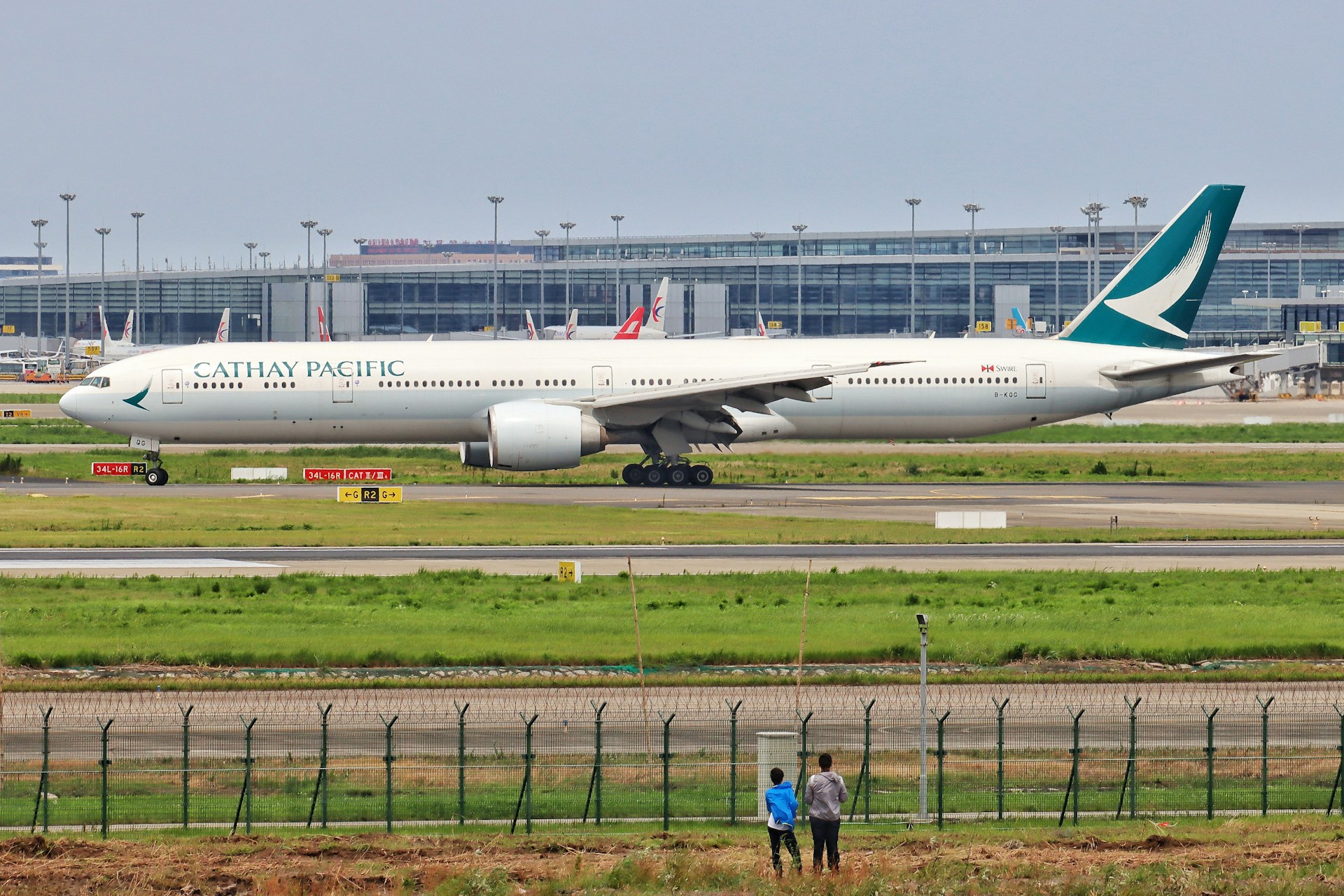 Wide angle shot of Cathay Pacific aircraft, two people in the foreground stood in front of a fence watching the aircraft.