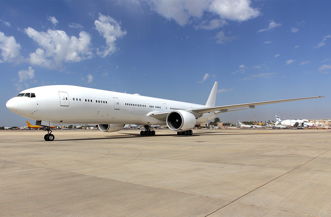 A plane is stationed on the ground in a taxiway, facing left, taking up the centre-left of the image into the centre. The background is sky blue and the foreground is sand coloured ground.