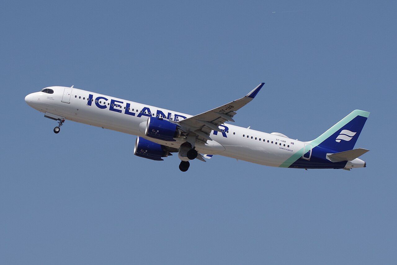 An Icelandair A321 flying under a blue sky. Its livery is mostly white with some dark and light blue. The logo is displayed on its side. The aircraft is departing from Schönefeld, Flughafen.